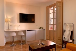 A modern living room with a flat-screen TV mounted on the wall, a wooden coffee table with remote controls, two bar stools, and a large window with a view.