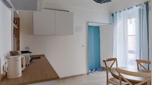 Una pequeña cocina con cafetera, lavamanos y armarios. Hay una puerta azul y una ventana con cortinas.
