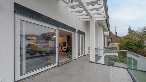 A modern building with large sliding glass doors leading to a spacious interior, a glass balcony with a metal railing, and a clear view of the surrounding area.