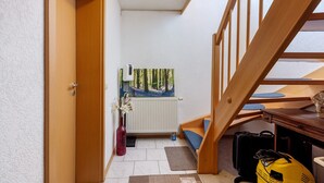A room with a wooden staircase, a radiator, a painting of a forest, a vase with flowers, and a suitcase.