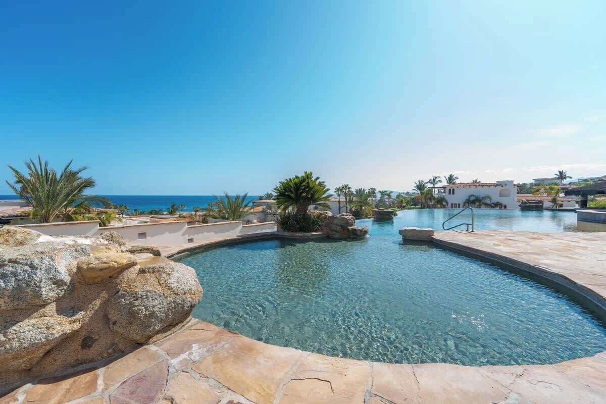 Una piscina con acqua cristallina, circondata da rocce e palme, con vista su una zona costiera e edifici sullo sfondo.