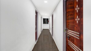 A long, narrow hallway with white walls and a dark floor, leading to a door with a decorative wooden panel. - Classic Laxmi Chowk Hinjewadi (Paud)