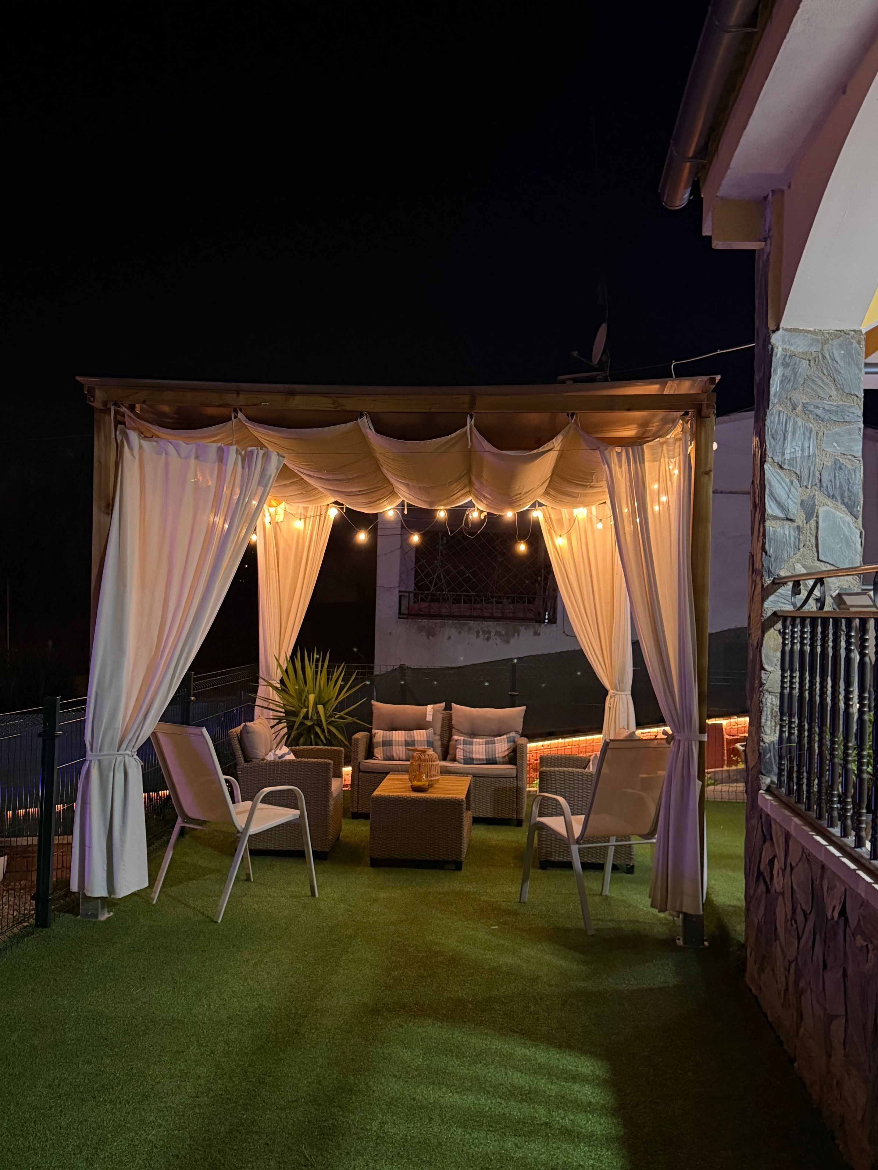 Un patio al aire libre con zona de estar cubierta, césped artificial y luces colgantes.