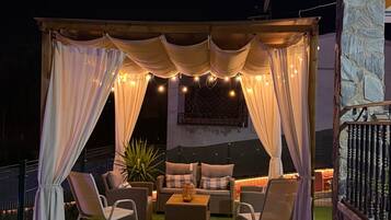 Un patio al aire libre con zona de estar cubierta, césped artificial y luces colgantes.
