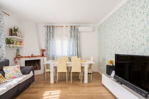 Una sala de estar con sofá, televisor, mesa de comedor, sillas, chimenea y plantas decorativas.