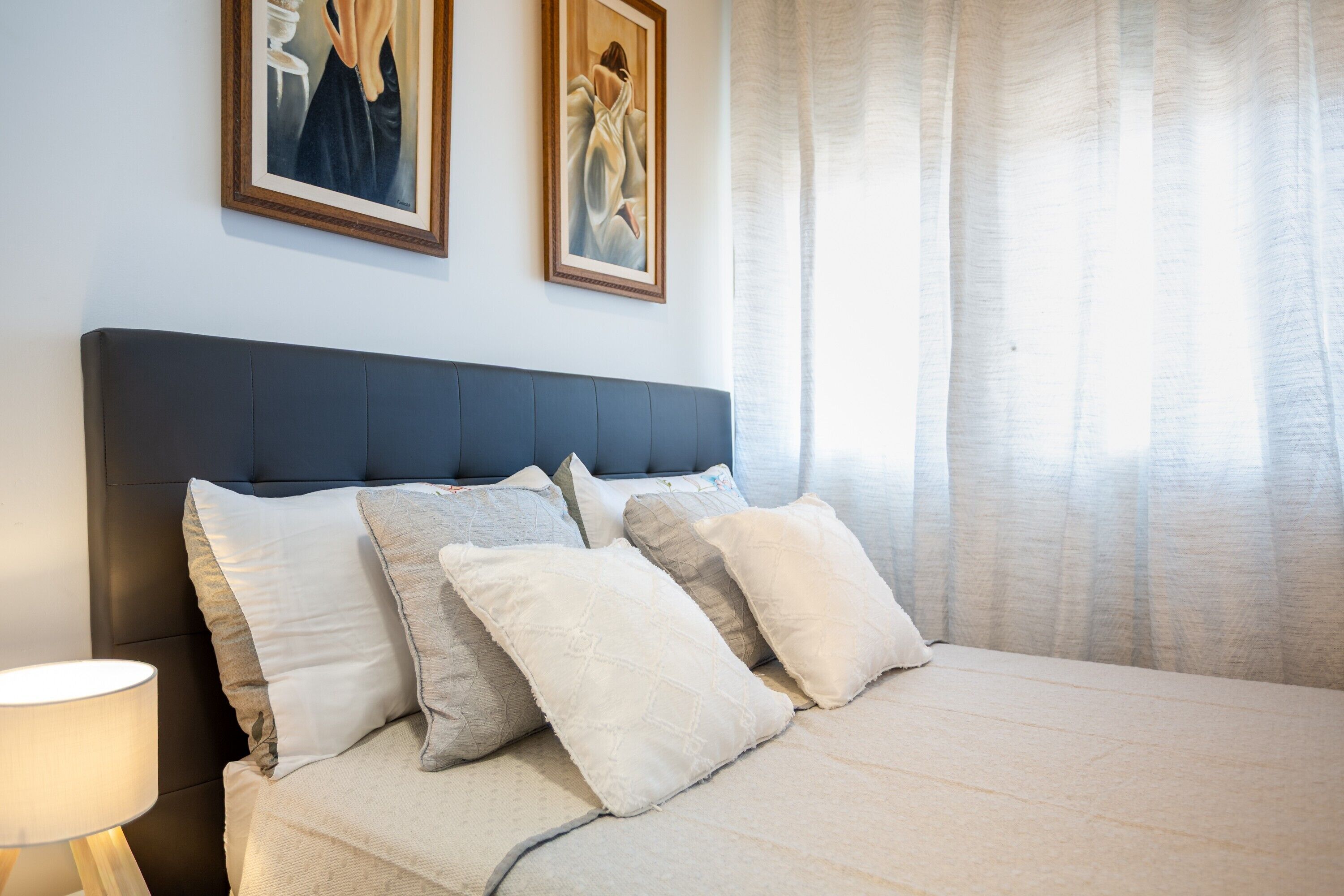 A bedroom with a dark headboard, multiple pillows, a bedside lamp, and two framed artworks on the wall.