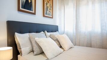 A bedroom with a dark headboard, multiple pillows, a bedside lamp, and two framed artworks on the wall.