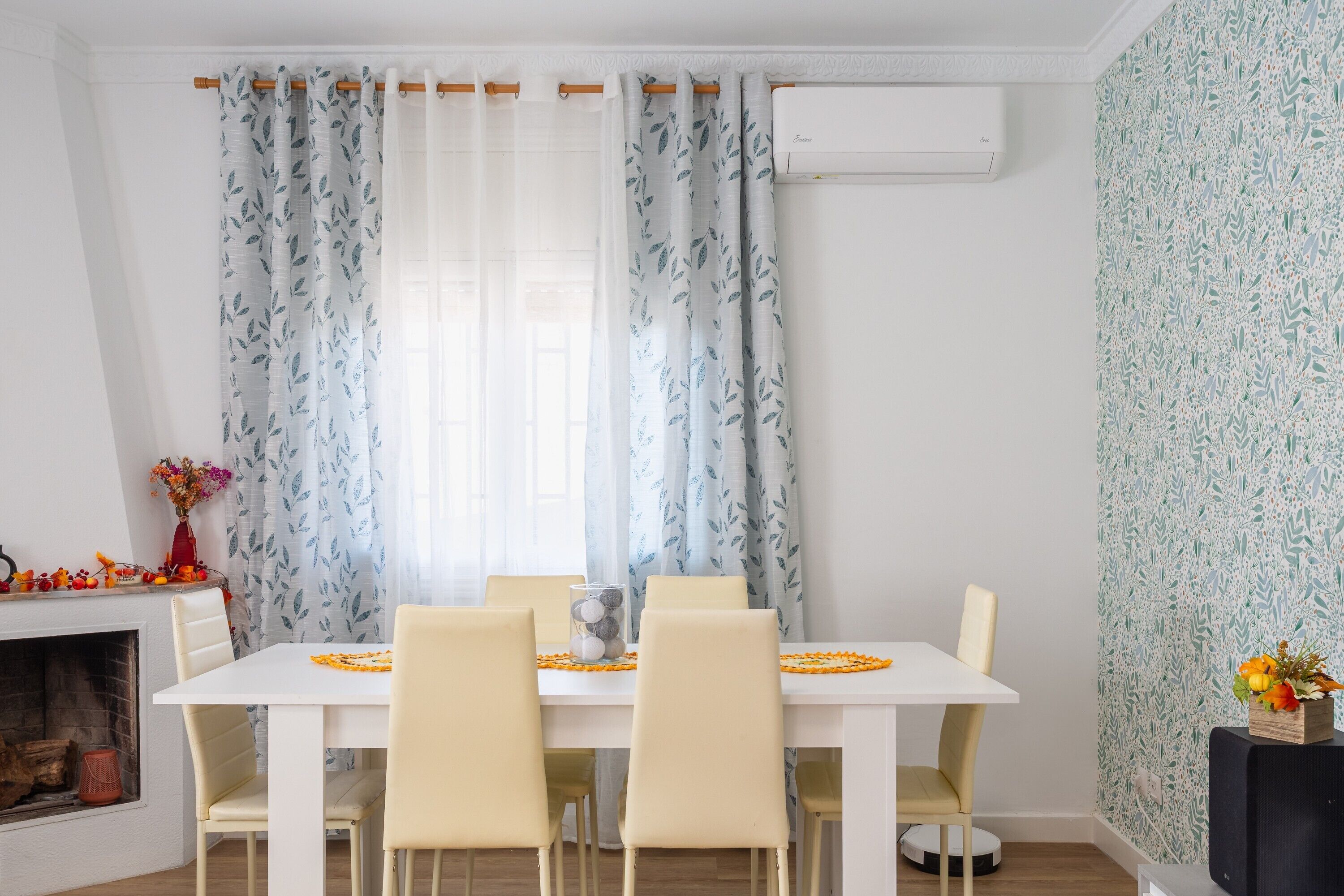 Un coin repas avec une table blanche, des chaises de couleur beige, une cheminée et des rideaux à motifs.