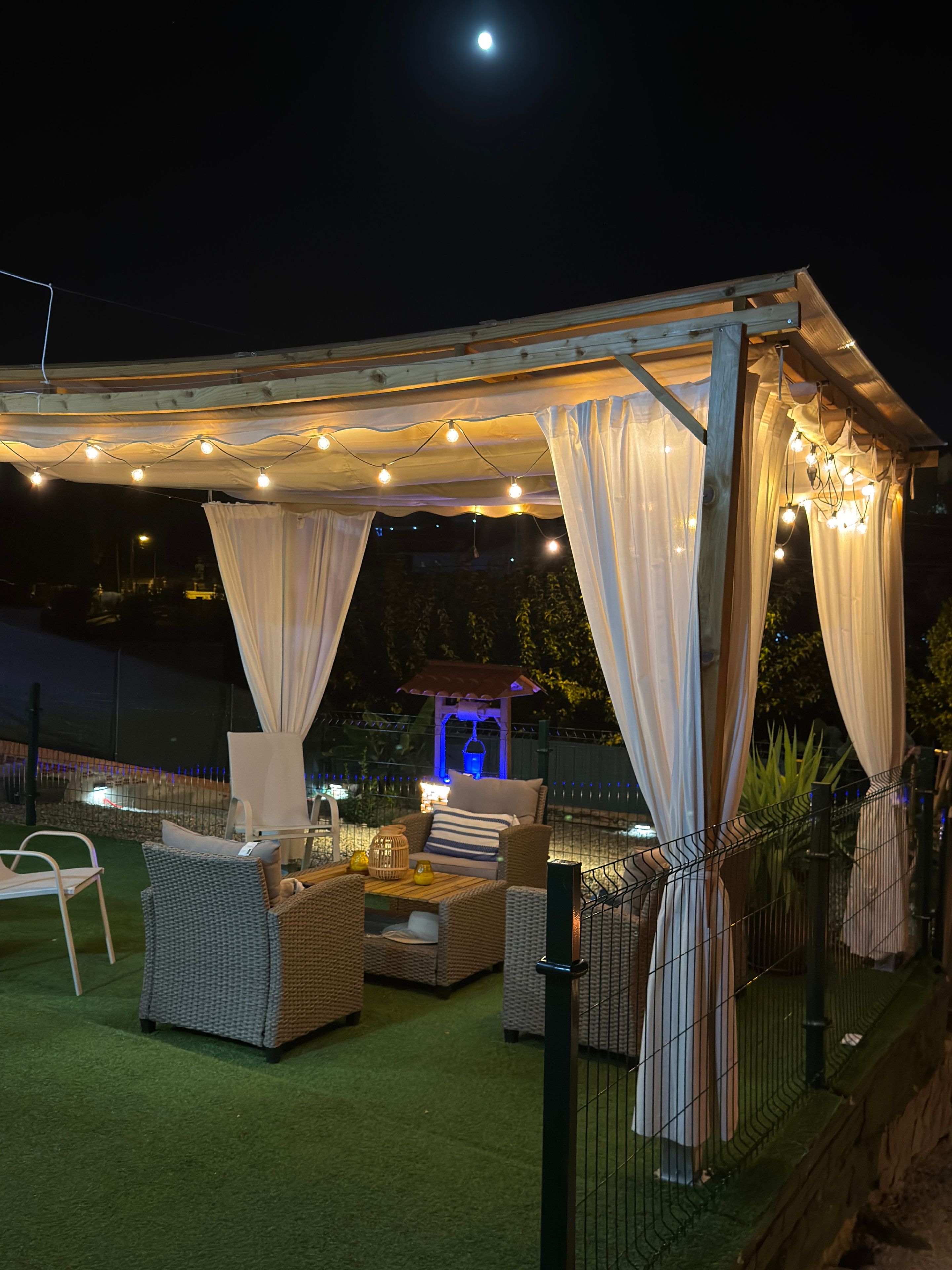 En udendørs patio med fletmøbler, en pergola med lyskæder og en lille oplyst bygning i baggrunden.