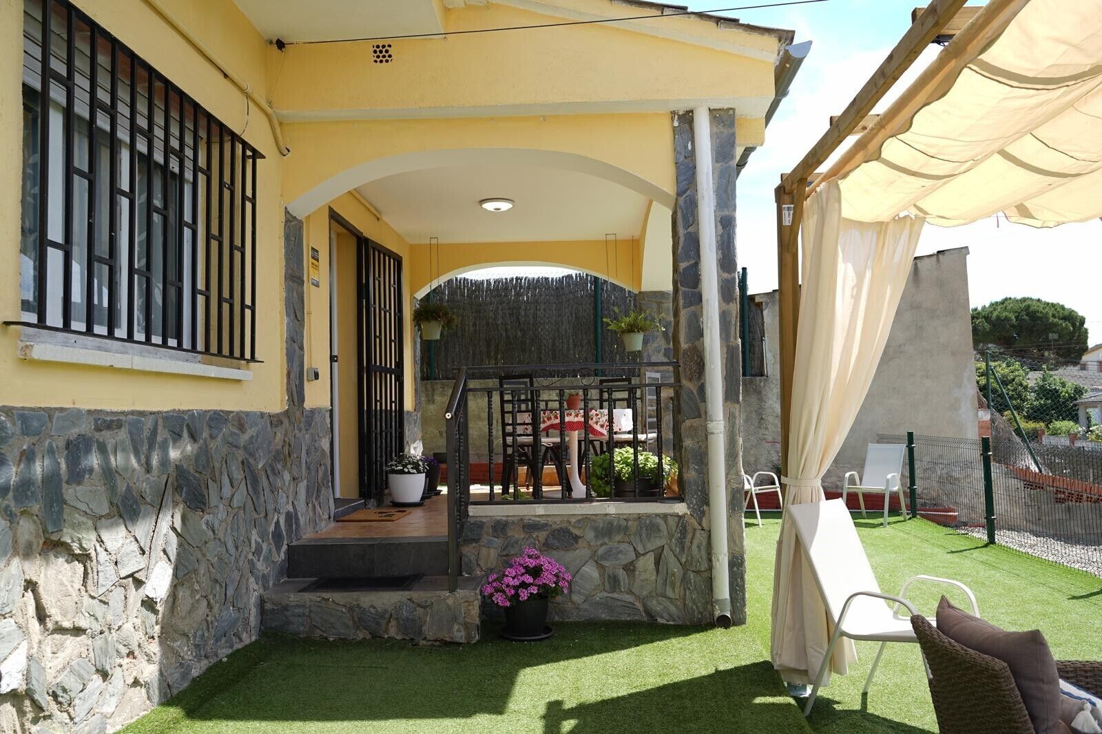 Una casa de fachada de piedra, con un balcón que tiene una mesa y sillas, y un patio cercado con césped artificial.