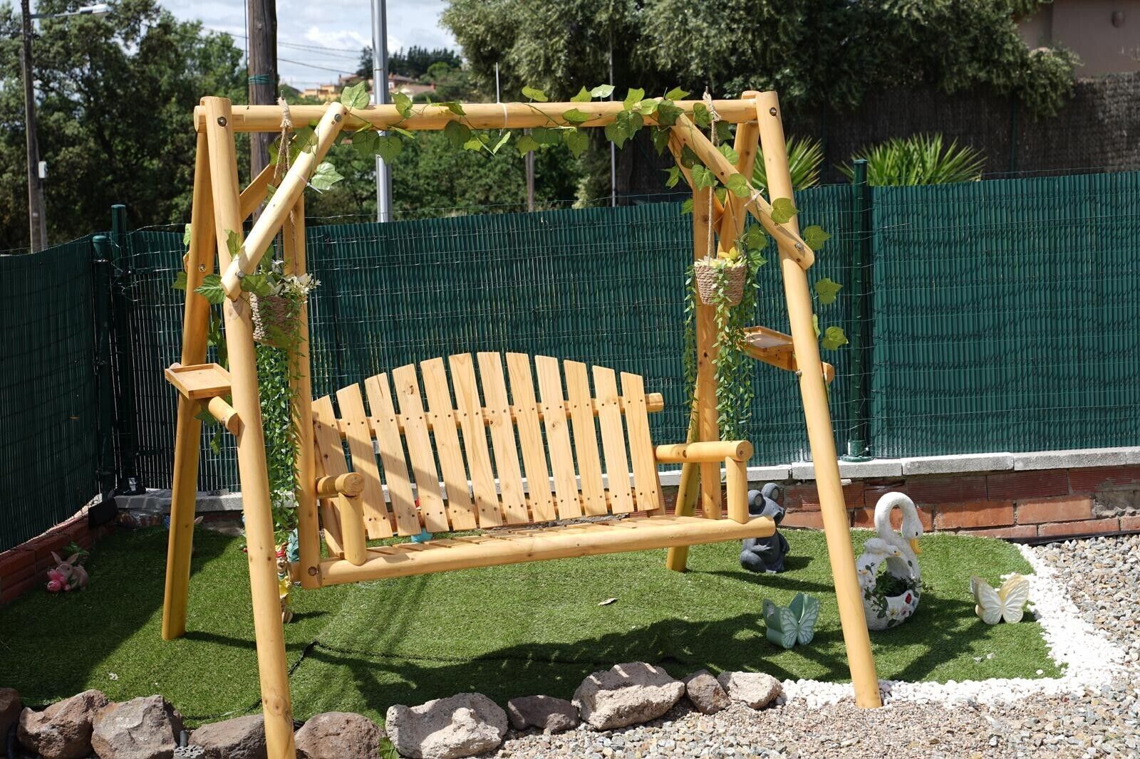 Un ensemble de balançoires en bois, agrémenté d’un banc, entouré d’une clôture verte, de rochers et de décorations de jardin.
