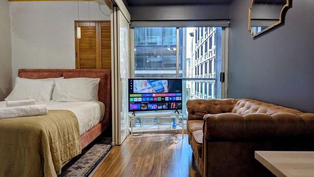 Quarto de hotel com uma cama, um sofá marrom, uma TV sobre um suporte e vista para prédios por uma janela grande.