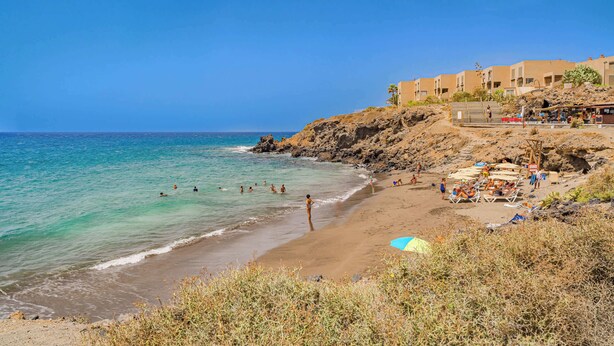 A beach with clear turquoise water, a sandy shore, and a building with multiple balconies overlooking the sea. - Sotavento Horizon Views Suite (La Tejita)