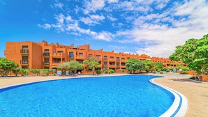 A large swimming pool with a clear blue water surface, surrounded by a tiled deck. In the background, there are multi-story buildings with balconies and windows. - Sotavento Horizon Views Suite (La Tejita)
