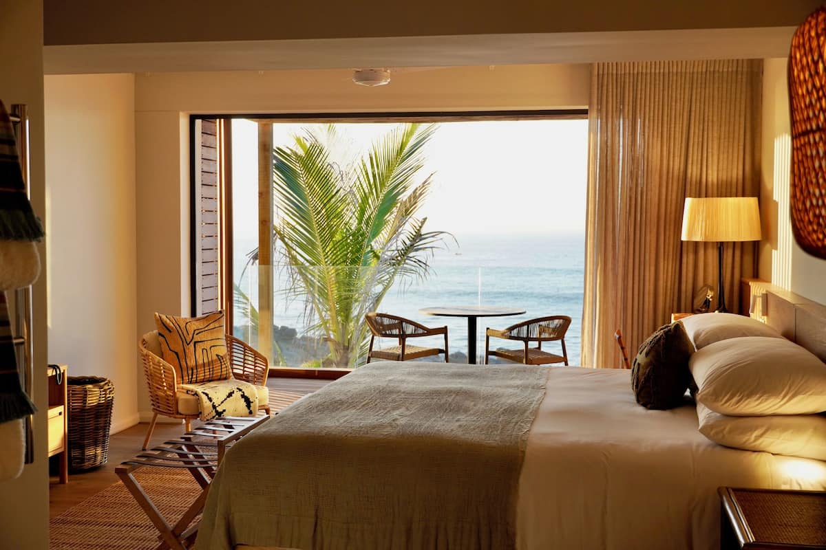 A hotel room with a large bed, a view of the ocean, a dining area, and a sitting area with a plant.