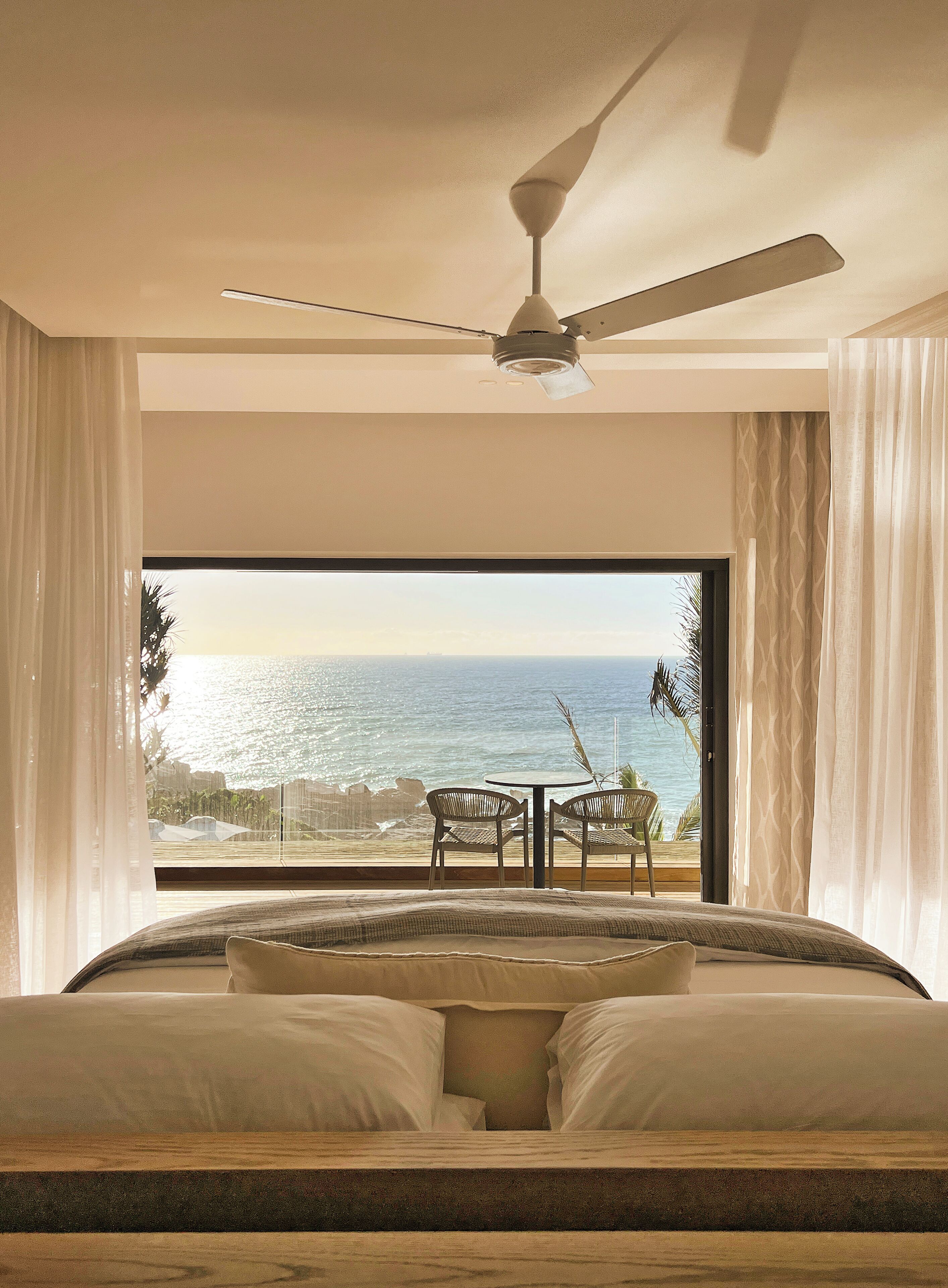 A bedroom with a large window, a ceiling fan, and a view of the ocean.