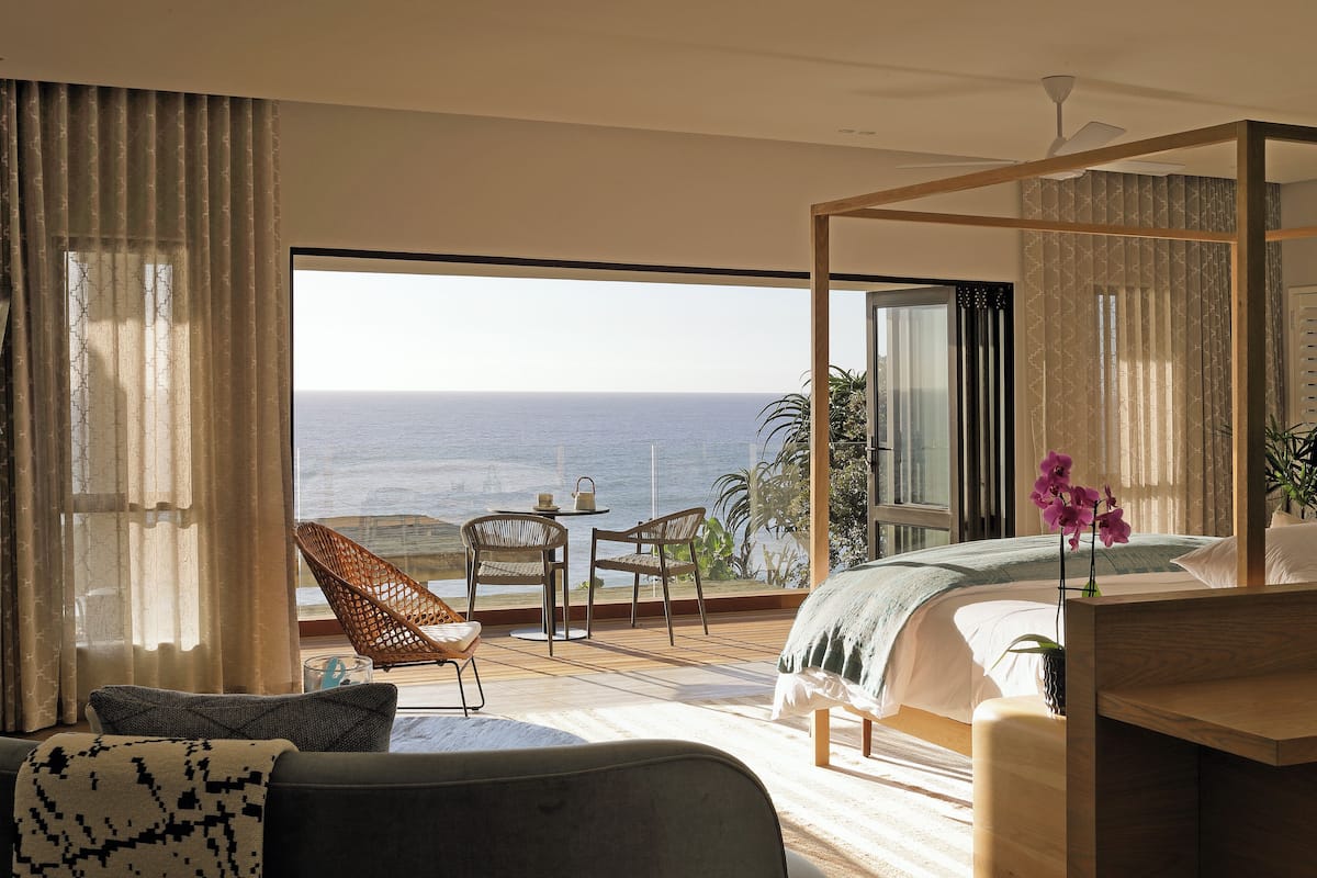 A bedroom with a large window overlooking the ocean, a bed, a bedside table, and a chair.