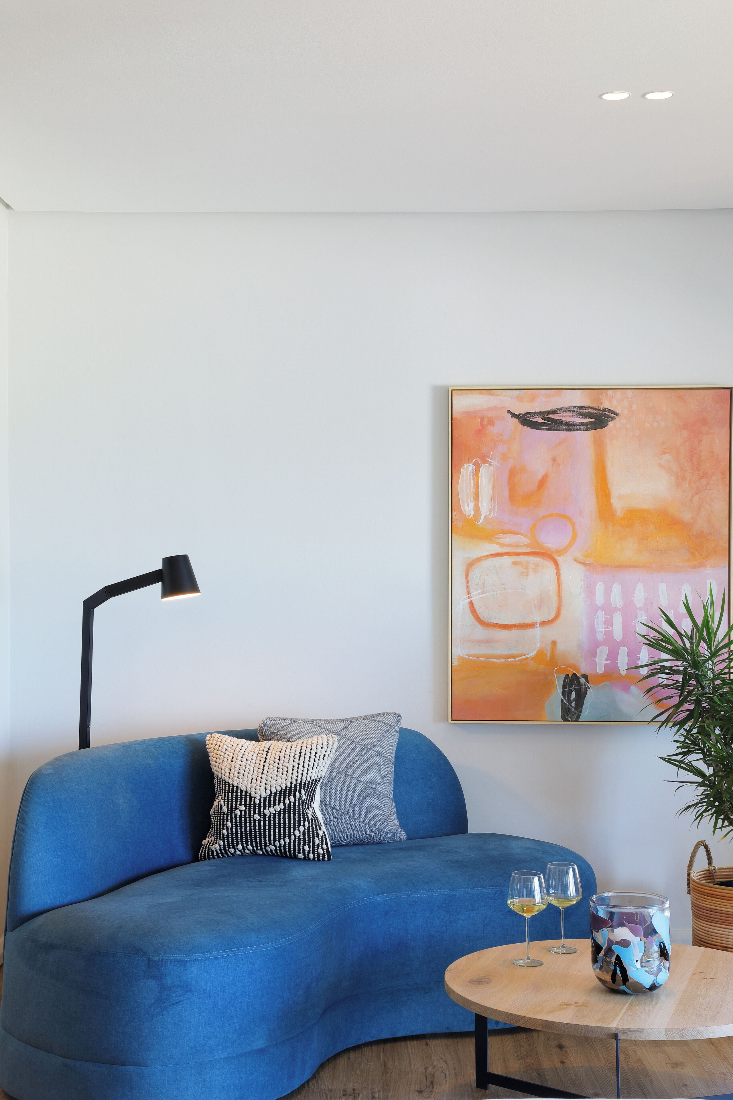 A blue sofa with a patterned pillow, a round wooden table with two glasses, a black floor lamp, and a painting on the wall.