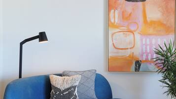 A blue sofa with a patterned pillow, a round wooden table with two glasses, a black floor lamp, and a painting on the wall.
