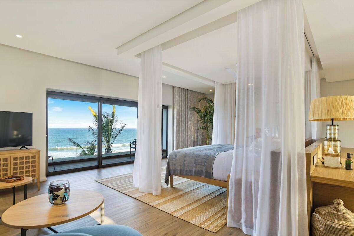 A modern hotel room with a large window offering a view of the ocean, a bed with a grey blanket, a bedside table with a lamp, and a television on a wooden cabinet.