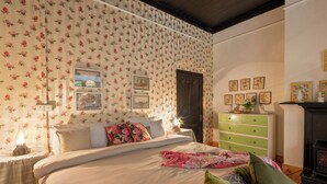 A bedroom with floral wallpaper, a bed with pillows, a dresser, and a fireplace.