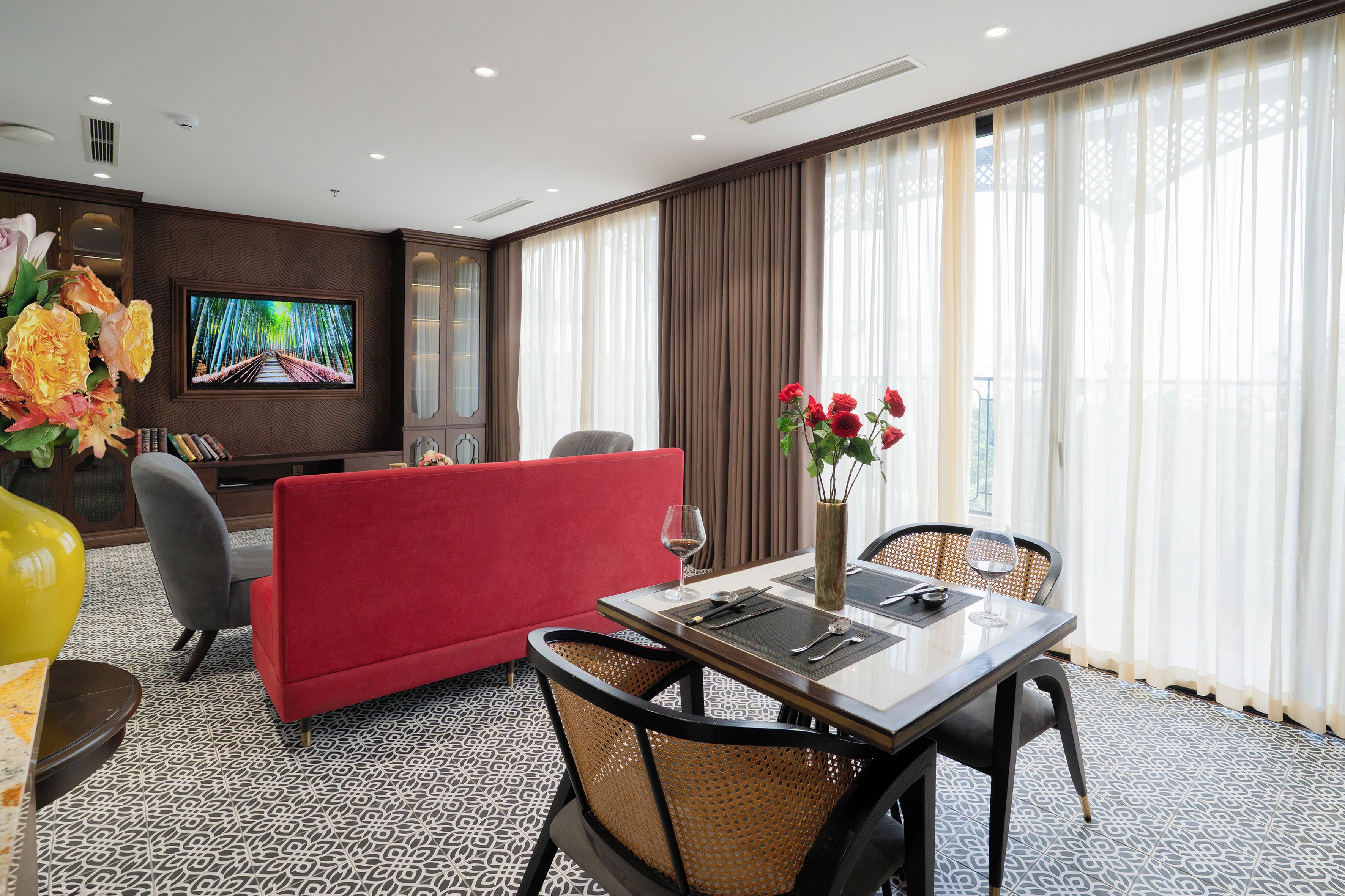 A hotel room with a red sofa, a dining table with chairs, a TV mounted on the wall, and a large window with curtains.