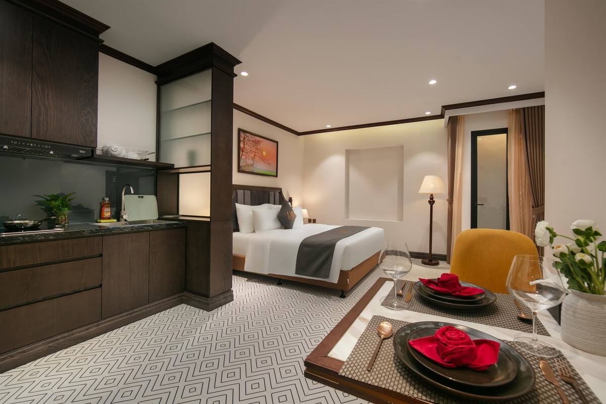 A hotel room with a bed, a dining area with a table set for two, a kitchen area, and a large window.
