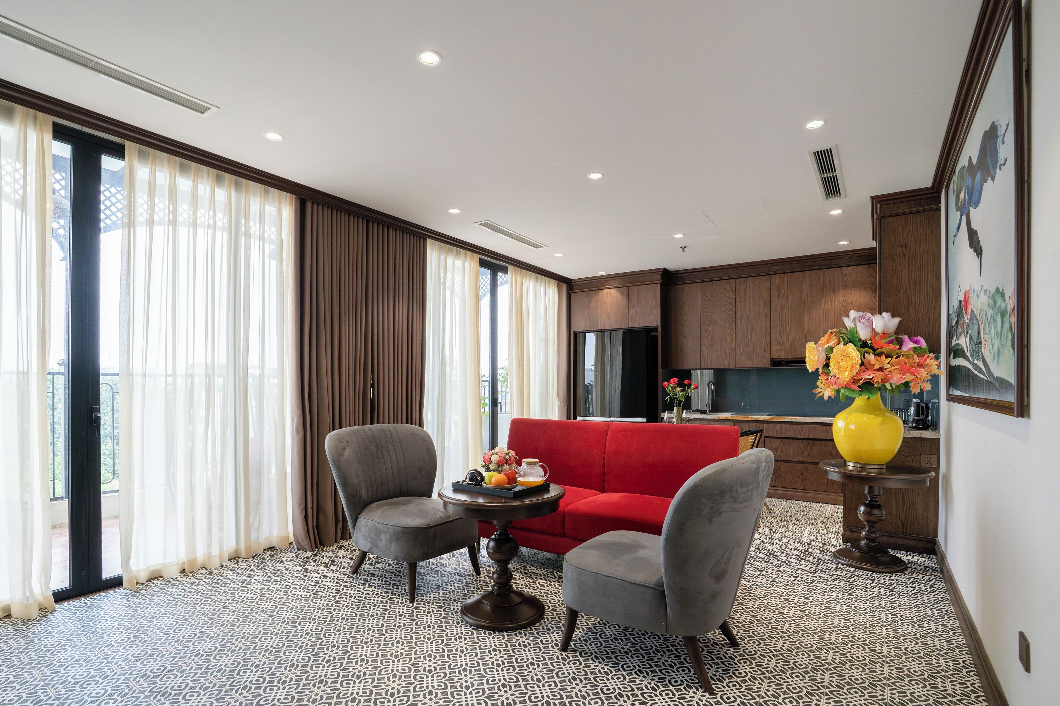 A modern hotel room with a red sofa, two armchairs, a small table with a vase of flowers, and a kitchen area in the background.