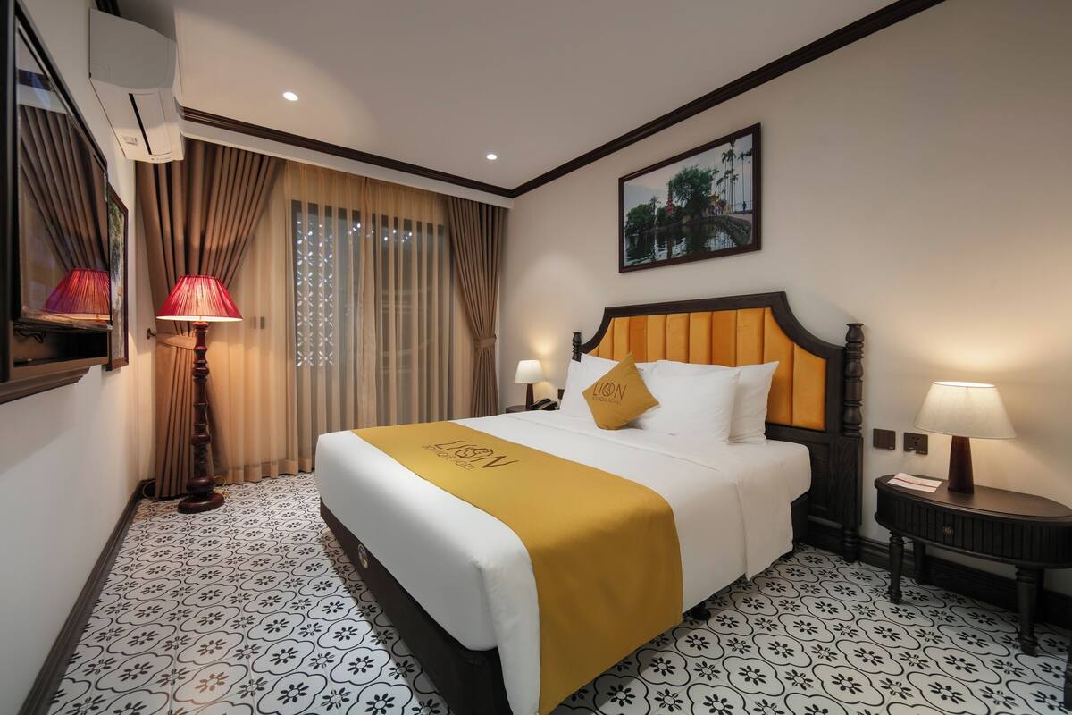 A hotel room with a large bed, a headboard, a nightstand, a lamp, a window with curtains, and a painting on the wall.