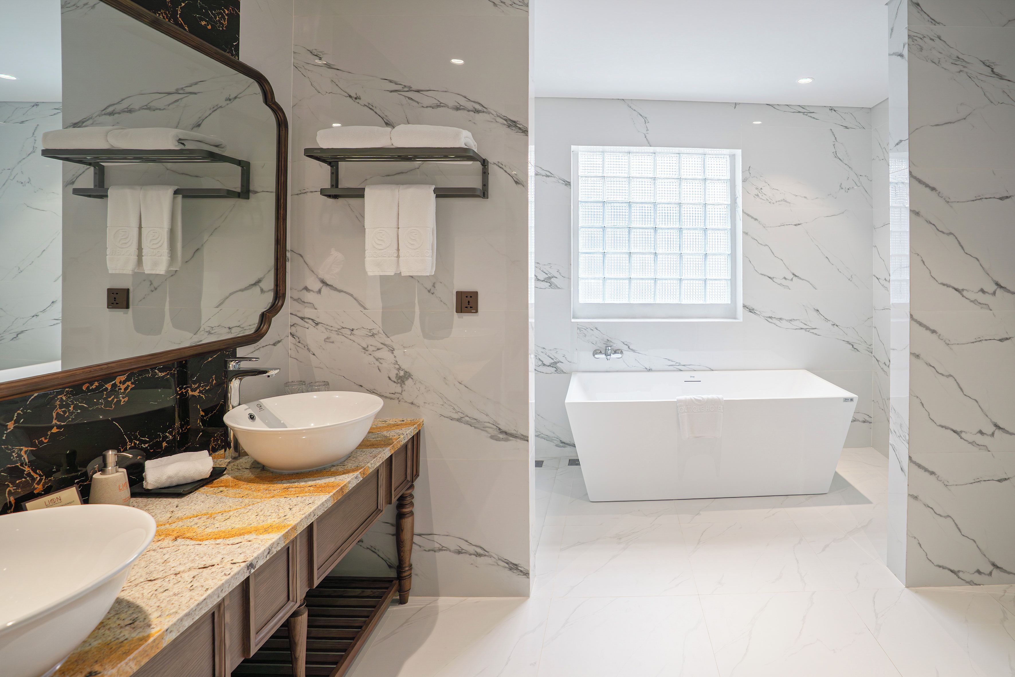 A modern bathroom with a marble wall, a large mirror, a sink, a bathtub, and a window with a view.