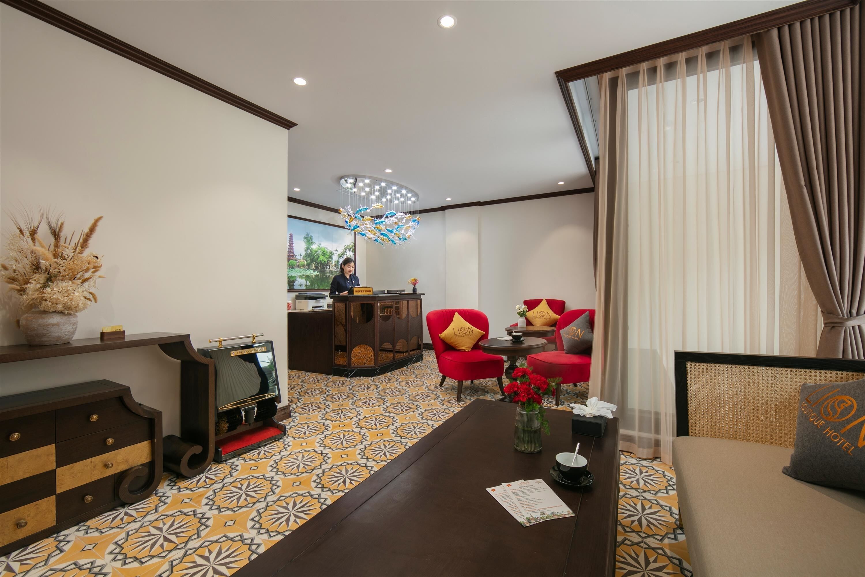 A hotel lobby with a reception desk, red and beige chairs, a patterned floor, and a decorative chandelier.