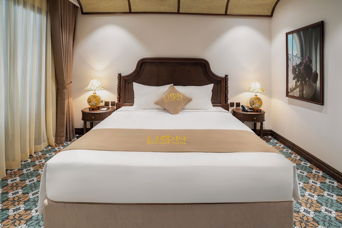 Une chambre d’hôtel avec un grand lit, deux tables de chevet avec des lampes, une tête de lit, un tableau accroché au mur et une couvre-lit ornée du logo de l’hôtel.