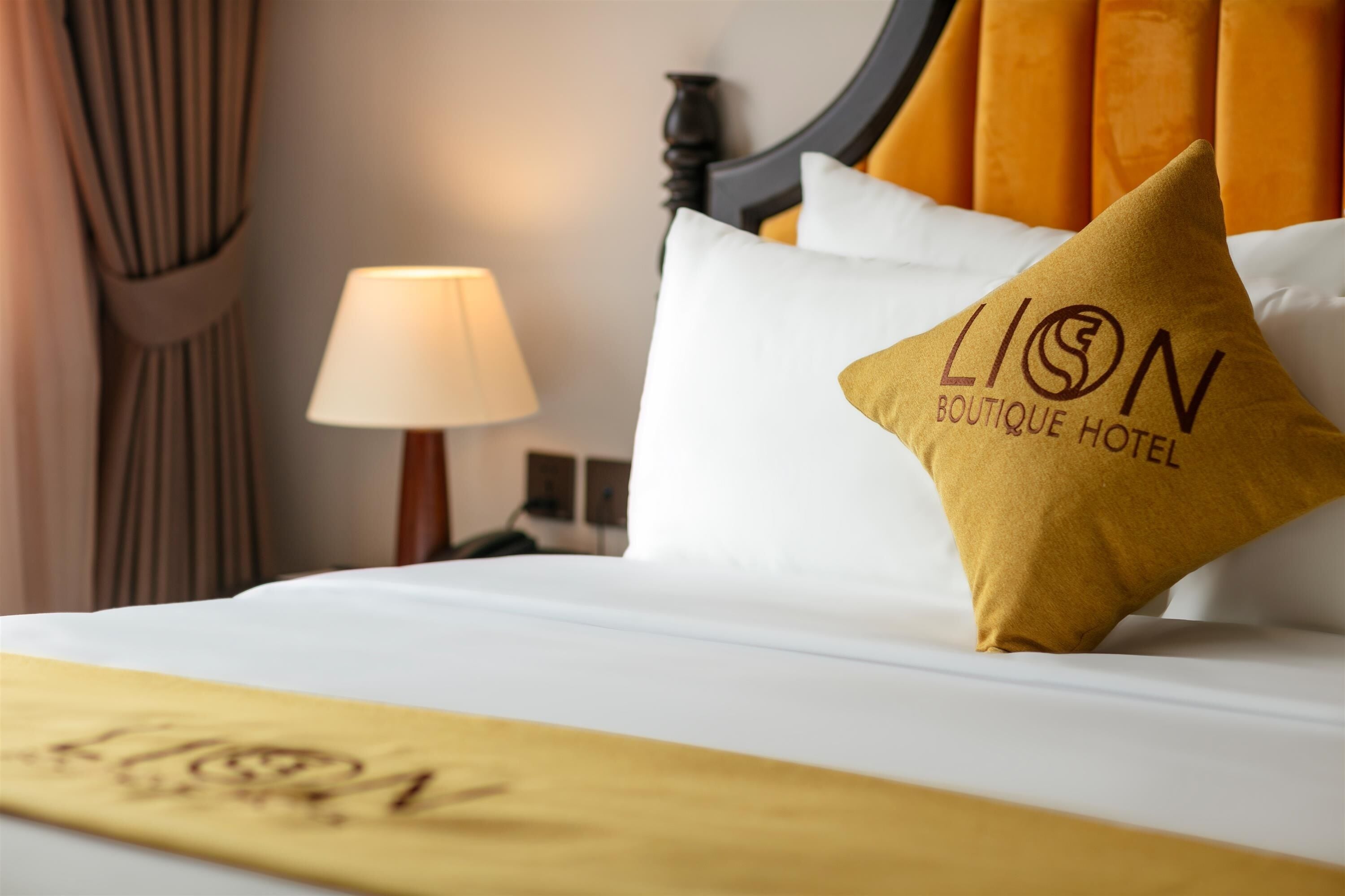 A hotel room with a bed, a pillow with the hotel's logo, a lamp, and a curtain.