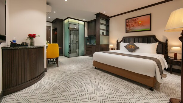 A hotel room with a large bed, a desk with a vase of flowers, a bathroom with a glass door, and a painting of a sunset on the wall. - Westlake Lion Boutique Studio & Spa (Hanoi)