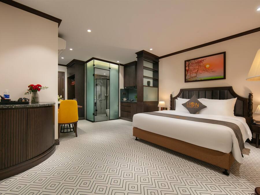 A hotel room with a large bed, a desk with a vase of flowers, a bathroom with a glass door, and a painting of a sunset on the wall.