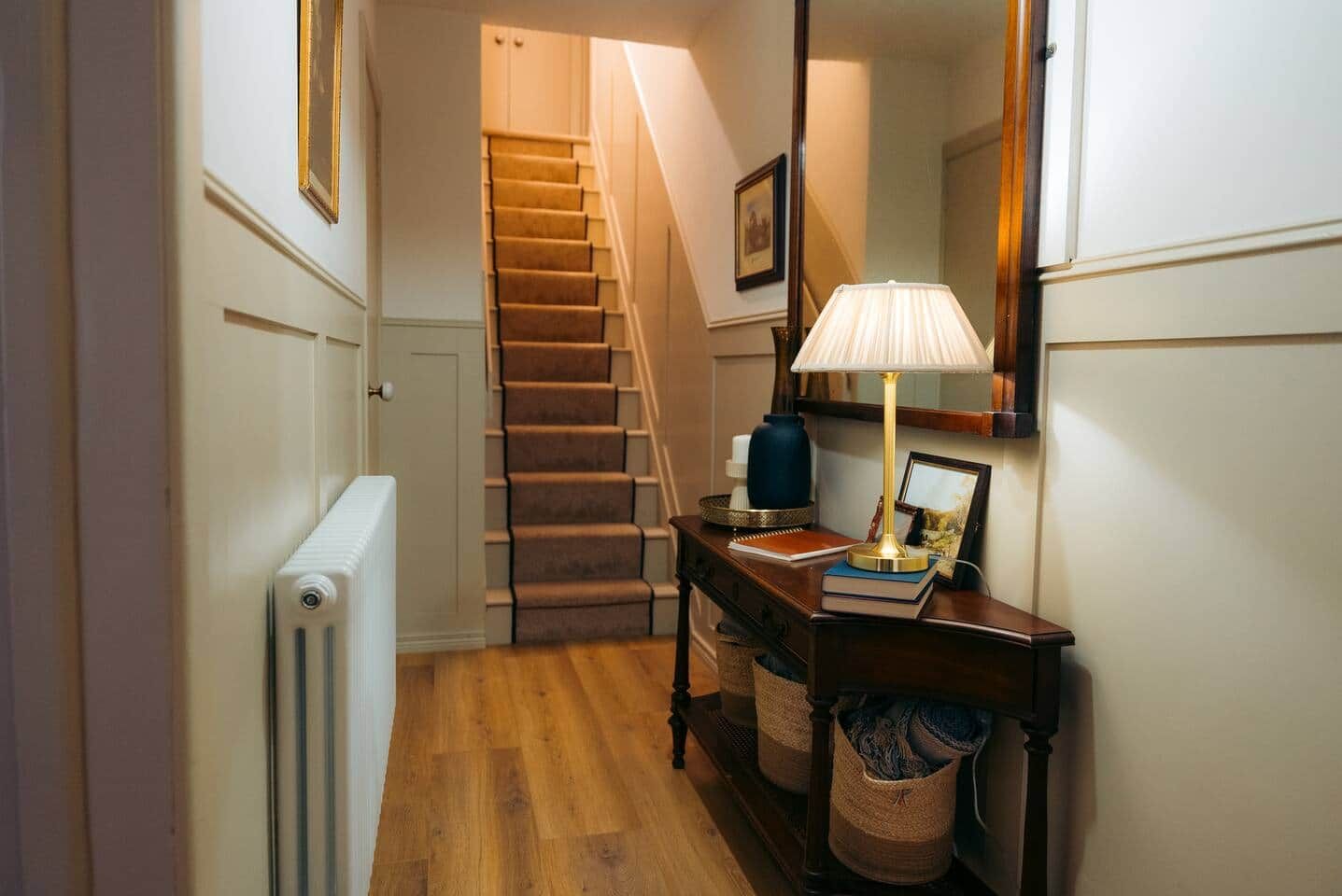 Een hal met een radiator, een houten vloer, een trap, een bureau met een lamp en een spiegel.