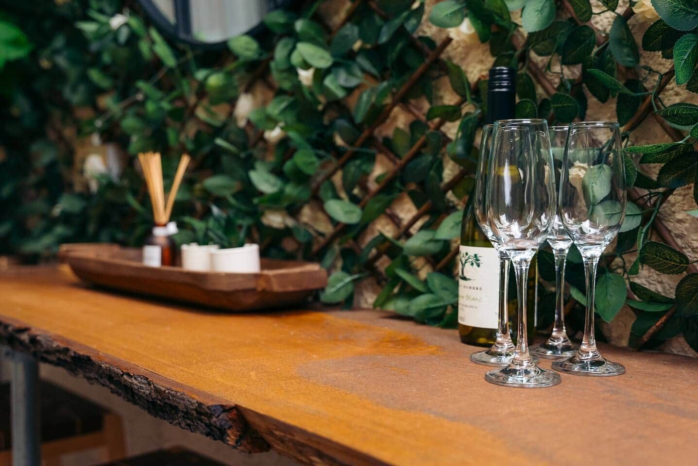 Balcão de madeira com três copos de vinho e uma garrafa, cercado por vegetação.