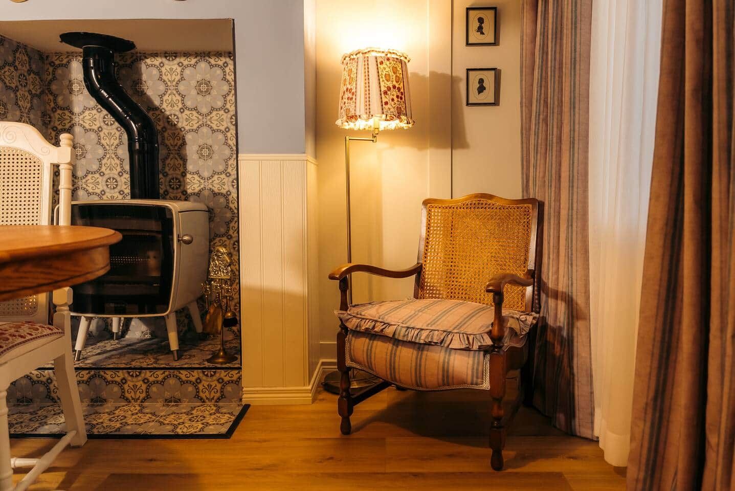 Une pièce avec un poêle ancien, une chaise en osier, un lampadaire et un papier peint à motifs.
