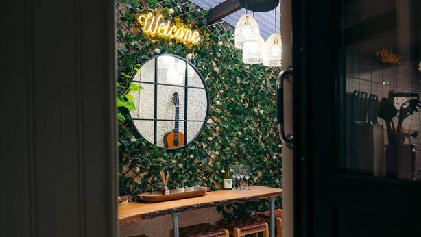 Zona de bar con una guitarra, un letrero de 'Welcome' y una pared cubierta de follaje.