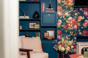 Una habitación con pared azul, papel tapiz floral, estantería, silla y un florero con flores.