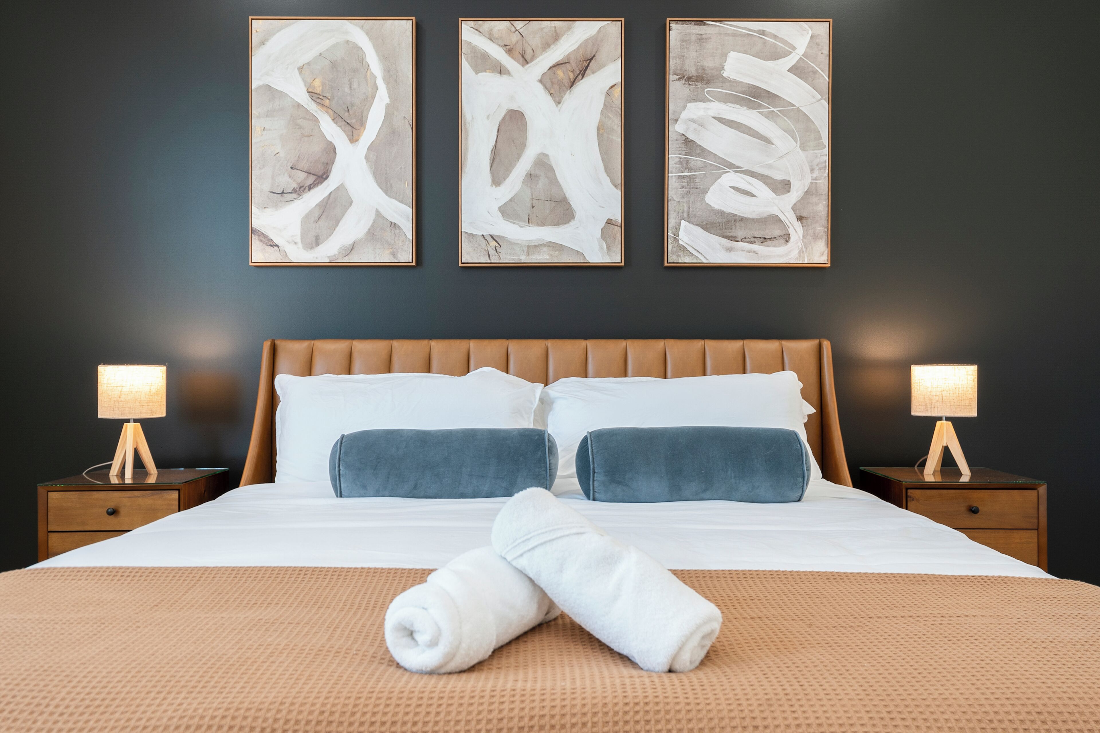 A neatly made bed with a brown headboard, two bedside tables with lamps, and three abstract paintings on the wall.