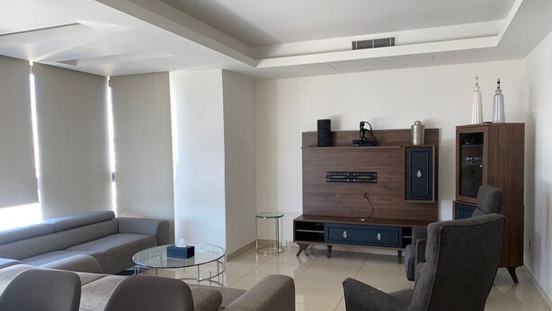 A modern living room with a sectional sofa, a glass coffee table, a dark armchair, a wooden entertainment unit, and a cabinet with decorative items. - Apartment in the heart of hazmieh (Hazmieh)
