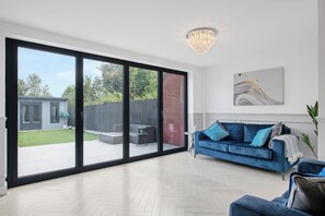 Een ruime woonkamer met een grote schuifpui naar een achtertuin, een blauwe bank en een moderne kroonluchter.