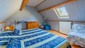 Une chambre avec un plafond en pente, un lit recouvert d’une couvre-lit à motifs, une commode en bois, un radiateur et une fenêtre donnant sur des arbres.