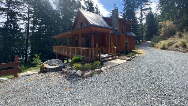 A wooden cabin with a porch, surrounded by trees and a gravel driveway.