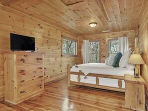 A wooden cabin room with a bed, a dresser, a television, and windows.