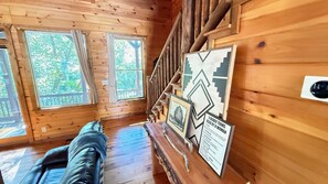 A cabin interior with a wooden staircase, a bed, and a wall with framed pictures and a sign.