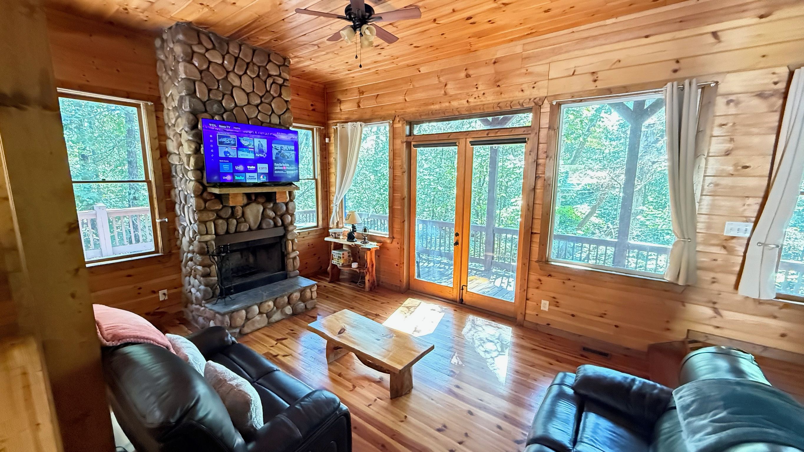 A wooden cabin interior with a stone fireplace, leather sofas, and large windows.