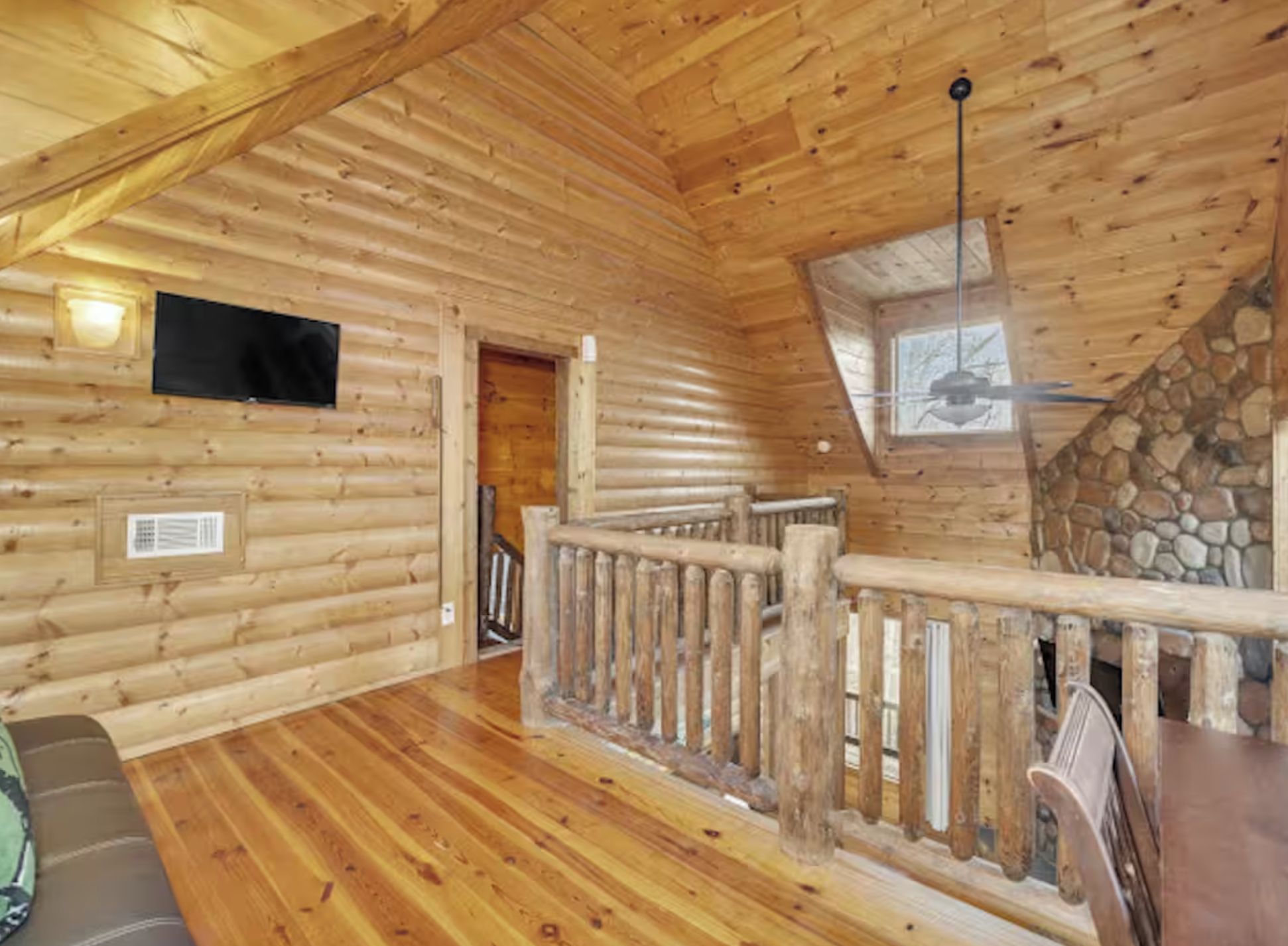 A log cabin interior with a wooden floor, a TV mounted on the wall, a staircase with a stone wall, and a wooden railing.