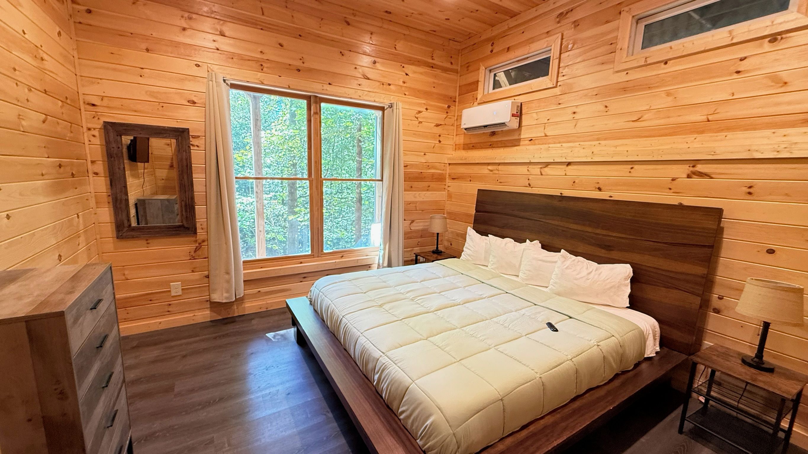 A wooden cabin room with a bed, a dresser, a window with curtains, and an air conditioning unit.